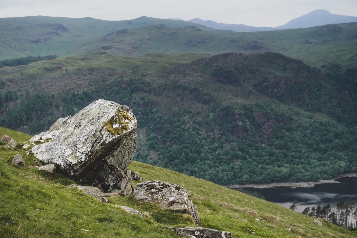A boulder on a grassy slope above a wooded valley and a lake