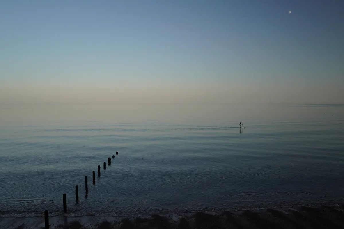 A calm sea at dusk with a line of wooden posts and a distant paddleboarder