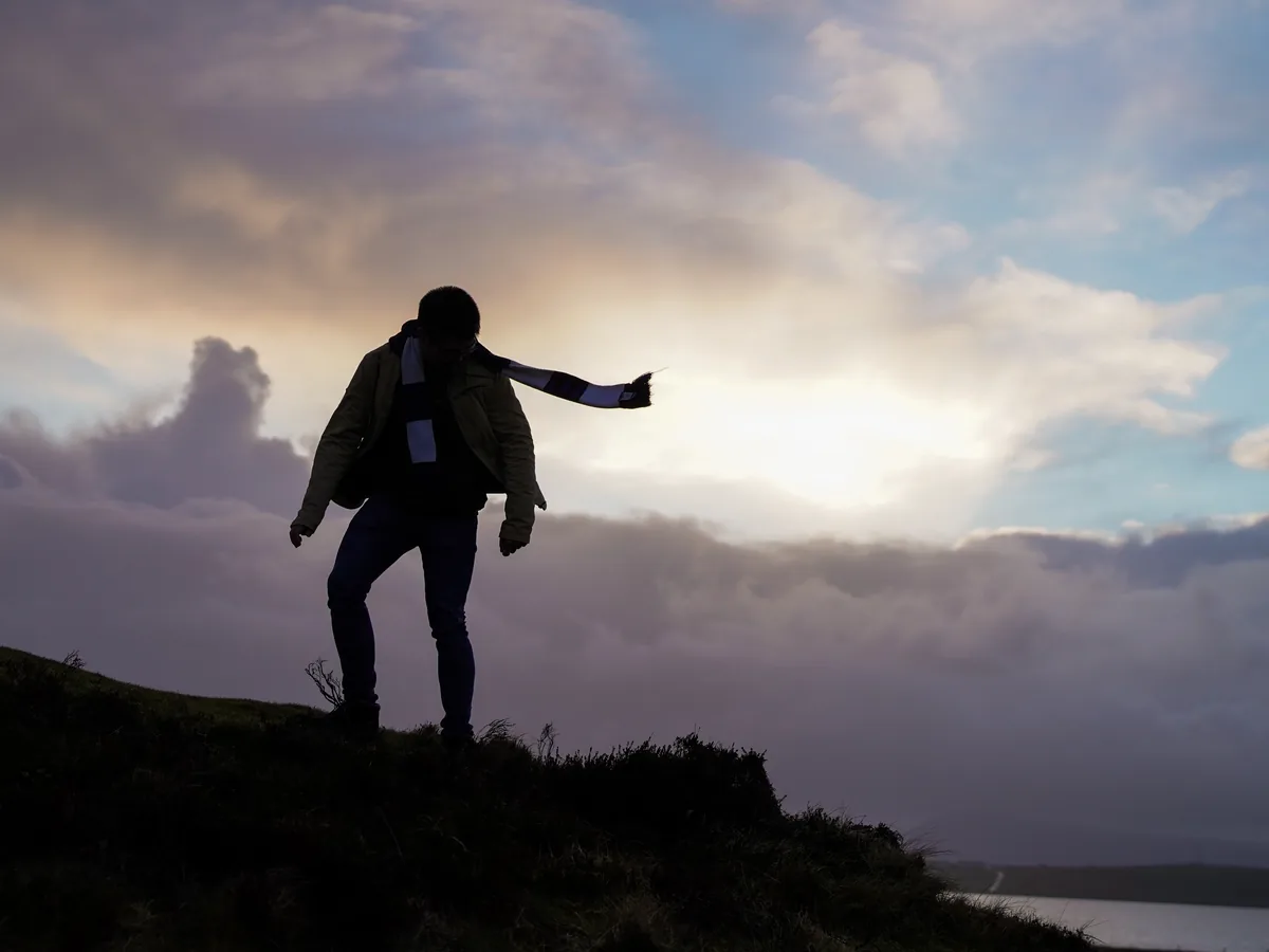 A figure in silhouette on a hilltop, one arm outstretched, against bright clouds