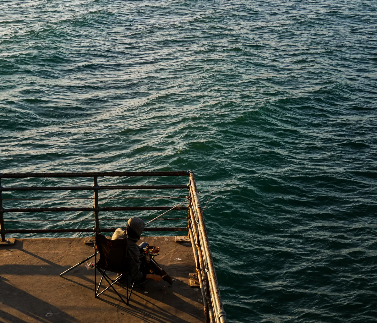 A person sitting at the corner of a pier above dark green waves