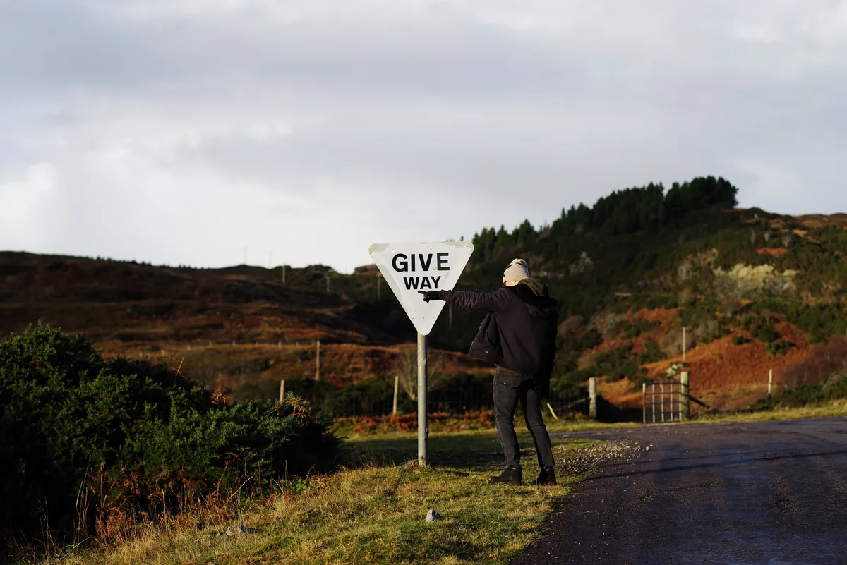 A person leaning on a Give Way sign beside a moorland road